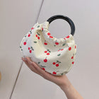 Cream canvas cherry print handbag with a black circular handle held by a hand.