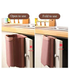 Foldable pink hanging kitchen trash can attached to a cabinet door for space-saving storage.