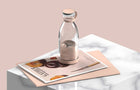 Clear glass juice bottle with a peach-colored lid and handle on a marble surface.